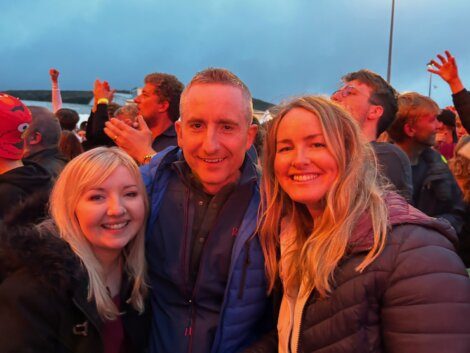 Three people are smiling at a crowded outdoor event. The two women are standing on either side of the man, all facing the camera. The crowd behind them appears to be celebrating or enjoying the event.