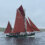 A green and black traditional sailing boat with brown sails, labeled LK 243, floats on calm waters with a few people onboard and land in the distant background.