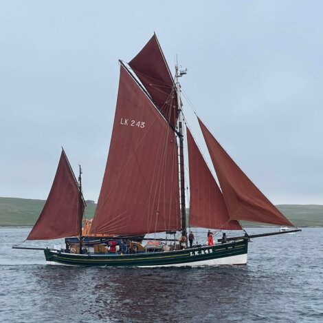 A green and black traditional sailing boat with brown sails, labeled LK 243, floats on calm waters with a few people onboard and land in the distant background.