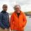 Two men stand outdoors on a rocky coastal landscape. One wears a blue jacket and cap, the other an orange puffy jacket. Both smile at the camera under a cloudy sky.