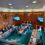 A group of people in formal attire sit around a U-shaped table in a wood-paneled room, conducting a meeting with some participants joining via video conference displayed on screens.