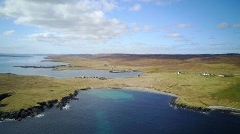 Aerial view of a coastal landscape with cliffs, green fields, scattered buildings, and a body of water under a blue sky with clouds.