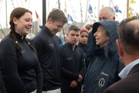 A woman in a raincoat is talking to a group of people in matching jackets. They appear to be outdoors near boats with various flags displayed.