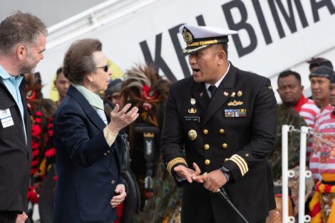 A woman in a green jacket converses with a uniformed naval officer while a man in a black jacket stands beside them. Several people with traditional attire are visible in the background.