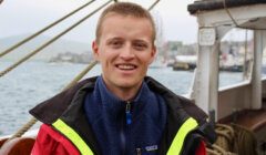 A person wearing a red and yellow jacket and a blue fleece stands on a boat. The background shows blurred water and harbor scenery.