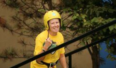A person wearing a yellow helmet and yellow shirt is smiling and standing outdoors near a railing, with trees in the background.
