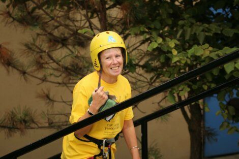 A person wearing a yellow helmet and yellow shirt is smiling and standing outdoors near a railing, with trees in the background.