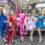 A group of five people in colorful and extravagant costumes pose confidently in front of a truck during a street event. The surrounding area includes shops and pedestrians.