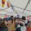 A group of people are gathering under a white tent decorated with colorful nautical flags, participating in a social event with food and drinks.