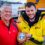 Two men stand in front of a lifeboat. The older man is in a red jacket, and the younger man, in a yellow RNLI jacket, holds a wooden plaque with a shield emblem.