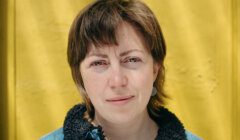 Woman with short brown hair and fair skin standing in front of a yellow background, wearing a blue jacket with a dark collar.