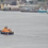 An orange tugboat guides a small sailboat near a large docked cruise ship in an industrial port area with various buildings and vessels in the background.