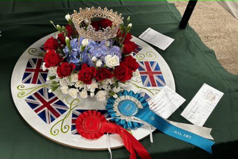 Floral arrangement with a crown, surrounded by Union Jack flags, a red and blue rosette, and an award certificate on a green surface.
