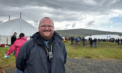 A smiling man in a dark jacket stands outdoors in front of a tent with a crowd of people in the background under a cloudy sky.