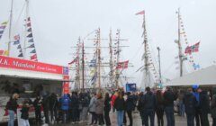 People are lined up at a catering truck on a cloudy day, with several tall ships adorned with flags in the background.