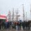People are lined up at a catering truck on a cloudy day, with several tall ships adorned with flags in the background.