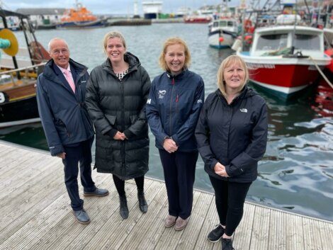 Four individuals stand side by side on a wooden dock with various boats moored in the background on a calm day.
