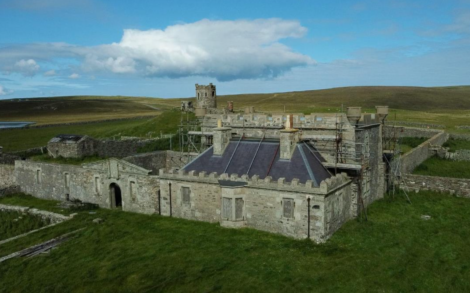 A stone building with crenellations and a central tower is surrounded by scaffolding, indicating restoration work. It sits in a large, grassy, rural area under a blue sky with clouds.