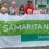 A diverse group of people standing outdoors holding a large banner that reads "SAMARITANS Shetland." They are in front of a gray building with a red fence on the left.