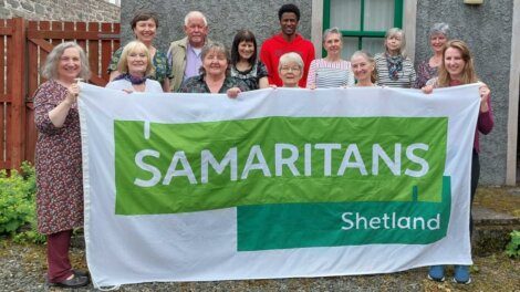 A diverse group of people standing outdoors holding a large banner that reads "SAMARITANS Shetland." They are in front of a gray building with a red fence on the left.