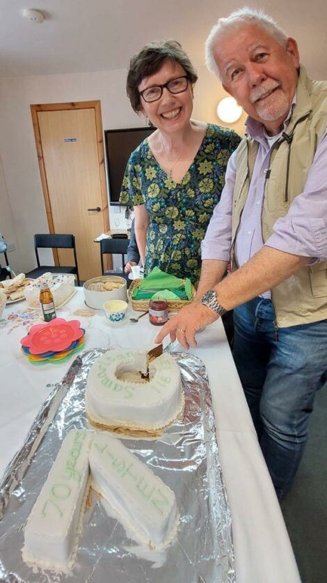 Two people stand smiling as one of them cuts into a decorated cake shaped like the letter "A." The table is set with various dishes and the background shows a closed door and a wall.