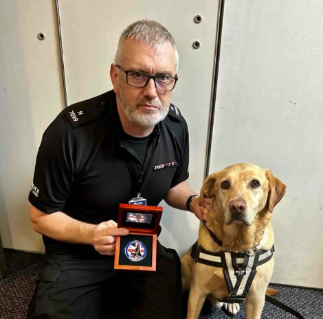 A man in a black uniform kneels beside a dog while holding a red box with a medal. The dog is wearing a harness.