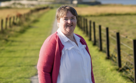 A smiling individual stands on a path flanked by fields and fences, wearing a white shirt and a pink cardigan.