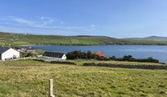 A scenic view of a coastal landscape with a large barn, a red house, and green hills under a clear blue sky.