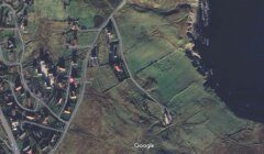 Satellite image displaying a coastal area with a blend of residential buildings, roads, and green fields next to an ocean.