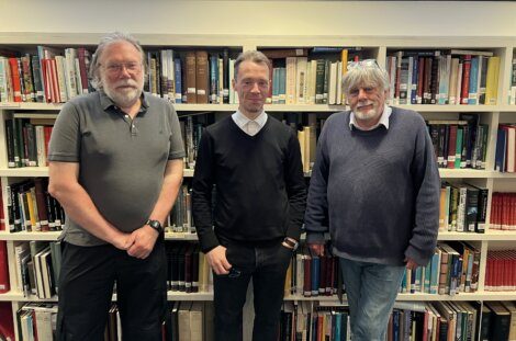 Three men are standing in front of a filled bookcase. The man in the middle wears a black sweater over a shirt, while the men on the sides wear casual shirts and sweaters.