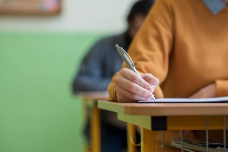 A person in an orange sweater is writing with a pen on paper at a desk in a classroom. Another person is blurred in the background.