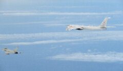 A military jet flies in close proximity to a larger reconnaissance aircraft over a cloud-filled sky.