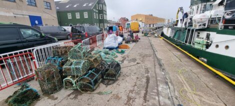 A dockside scene with fishing gear, including lobster pots and ropes, organized near a vessel. People are working in the background, and vehicles are parked nearby.