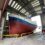 A large blue and white boat inside a covered dry dock with its hull exposed above yellow railings. Two people in red coveralls are visible on the right side of the image.