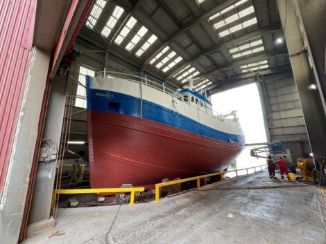 A large blue and white boat inside a covered dry dock with its hull exposed above yellow railings. Two people in red coveralls are visible on the right side of the image.