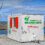 A white energy-efficient welfare unit from Ness Engineering Ltd on a gravel surface with wind turbines and a body of water in the background under a partly cloudy sky.