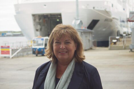 A woman with shoulder-length hair is standing in front of a large ship in a port area. She is smiling and wearing a dark jacket and light-colored scarf.