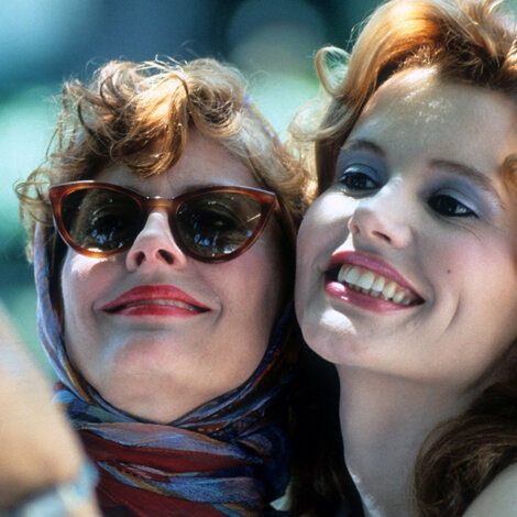 Two women smiling, one with sunglasses and a headscarf, the other with loose wavy hair. They are close together, appearing happy and relaxed.