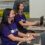 Two women wearing headsets work at desks with computers. They are both smiling and wearing purple shirts with an unreadable white logo. A scenic landscape image is in the background.