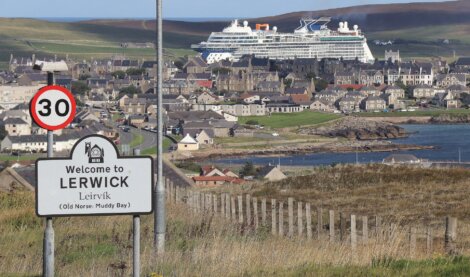 A road sign welcomes visitors to Lerwick, with a large cruise ship docked in the background and the town spread out along the coastline.