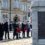 People stand in front of a memorial monument holding wreaths during a remembrance ceremony, with a building and military personnel in the background.