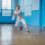 Person in white attire and black shoes performing a dynamic dance move in a spacious room with blue walls and windows.