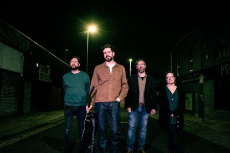 Four people stand in a dimly lit street at night. One person is holding a guitar. They all have neutral expressions and are facing the camera. The streetlights create a moody atmosphere.