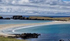 A serene coastal landscape featuring a sandy bay with clear blue waters, flanked by rocky outcrops under a partly cloudy sky.