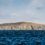 A large hill with rocky cliffs slopes gently into a blue ocean under a partly cloudy sky.