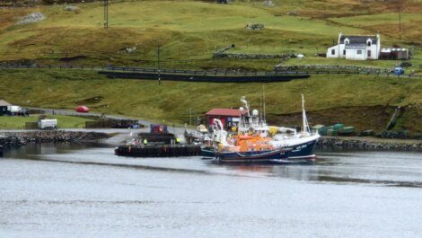 A fishing boat moored at a pier near a small white house with grassy hills in the background.