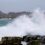 Waves crashing waves on rocks by a town with niagara falls in the background.