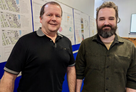 Two men standing in front of a board with papers on it.