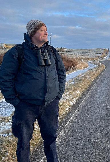 A person in winter clothing with binoculars hanging around the neck standing on a roadside, with snow on the ground and a clear sky overhead.