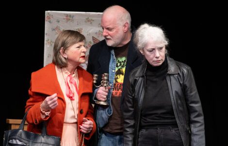 Three actors on stage performing a scene with one of them holding a bell and a discussion appearing to take place.